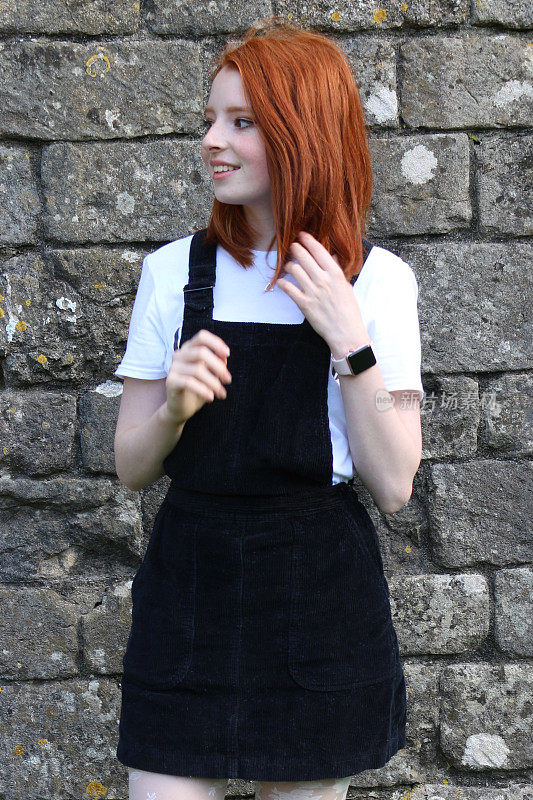 照片中，一个14 / 15岁的红发少女，皮肤苍白，脸上有雀斑，站在一堵长满青苔的旧石墙前，穿着一件时髦休闲的黑色罗纹灯芯绒围裙裙，梳着波浪发型